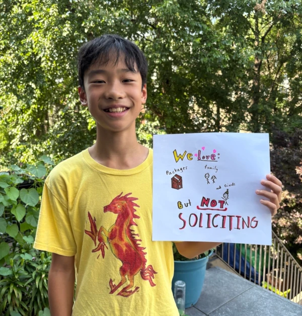 A smiling boy holding up a sign he drew, wearing a T-shirt with a dragon drawing on it